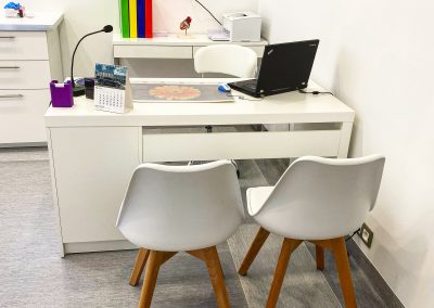 Białe biurko, na którym stoi laptop, lampka i kalendarz. Za biurkiem szafki i krzesło biurowe. Z przodu białe krzesła z drewnianymi nogami.